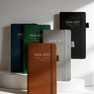 May include: A collection of 2026-2027 weekly and monthly planners in various colors, including black, navy, green, gray, and brown. Each planner features a matching elastic band closure and the text "2026-2027 Weekly & Monthly Planner" in gold lettering.
