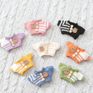 Puede incluir: Una colección de pequeños suéteres tejidos a mano en varios colores, incluyendo rosa, morado, amarillo, azul, verde, negro y naranja. Cada suéter tiene cierre de botones y un botón de madera con forma de rana. Los suéteres tienen diseños a rayas y lisos.