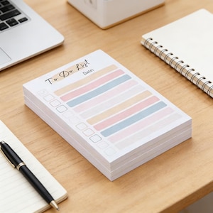 Könnte beinhalten: Ein Stapel pastellfarbener To-Do-Listen-Notizblöcke. Jeder Notizblock hat eine Kopfzeile mit den Worten "To Do List" und "Date:" und eine Reihe von Kontrollkästchen und horizontalen Streifen in Pastellfarben. Ein Stift und ein Notizbuch befinden sich im Hintergrund.