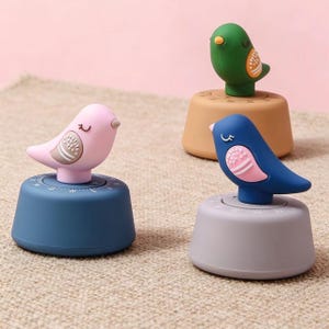 May include: Three bird-shaped kitchen timers in pink, green, and blue. Each timer has a round base with a numbered dial and a bird figure on top. The birds have white and pink accents. The timers are on a neutral surface.
