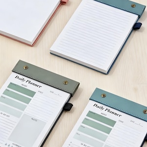 May include: Four daily planner notepads with leather covers in pink, teal, and green. Each notepad has a white paper interior and is labeled "Daily Planner". The notepads are on a light wood surface.