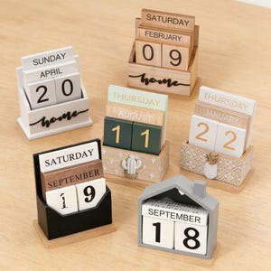 Pode incluir: Uma coleção de calendários perpétuos de madeira em várias formas e cores. Cada calendário mostra o dia, o mês e a data com blocos intercambiáveis. Os calendários são em branco, preto e tons de madeira natural, com elementos decorativos.