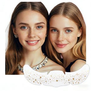 Op de afbeelding: Twee vrouwen met een lichte huid en sproeten, die sieraden dragen, lachen naar de camera. Een wit cosmetisch product met gouden sterren en stippen staat op de voorgrond. Het product is ontworpen om sproeten na te bootsen.