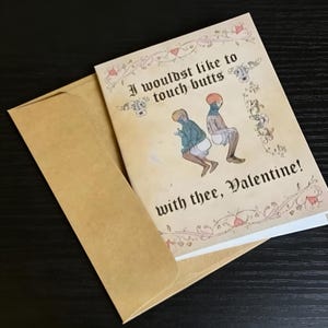 May include: A Valentine's Day card with a medieval illustration of two figures with the text "I wouldst like to touch butts with thee, Valentine!" The card is inside a brown envelope, set against a dark surface.