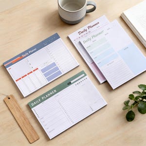 Puede incluir: Varios blocs de notas de planificación diaria con la inscripción "Daily Planner". Los blocs de notas tienen secciones para la programación, listas de tareas y notas. Un bloc de notas tiene un marcapáginas de madera al lado. Una taza y un libro también están en la imagen.