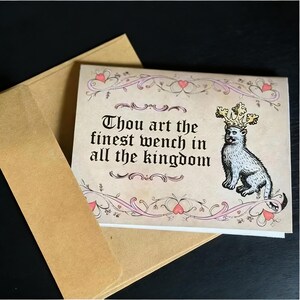 May include: Greeting card with a medieval theme. The card features a drawing of a cat wearing a crown, with the text "Thou art the finest wench in all the kingdom." The card is inside a brown envelope.