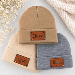 May include: Three knit beanies in tan, beige, and grey, each featuring a rectangular brown faux leather patch. The patches are secured with four metal rivets and are engraved with the names Clark, Ezra, and Flynn in a black script font.