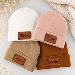 May include: Four knit beanies in white, light pink, tan, and brown. Each beanie features a rectangular brown faux leather patch attached with four metal rivets. The patches are engraved with the names Blake, Cody, Oliver, and Doyle respectively.