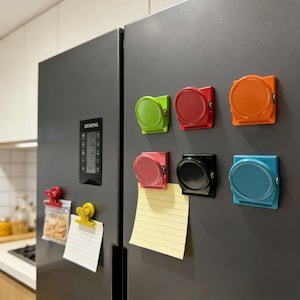 May include: A collection of colorful magnetic clips in green, red, orange, pink, black, and blue, attached to a gray refrigerator. Some clips hold notes and a bag of snacks. The clips have a circular design.