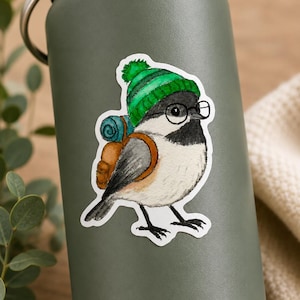 Peut inclure: Un autocollant représentant une illustration à l'aquarelle d'un petit oiseau portant un bonnet en tricot vert avec un pompon, des lunettes rondes noires et un sac à dos marron avec un sac de couchage bleu, collé sur une gourde vert olive.