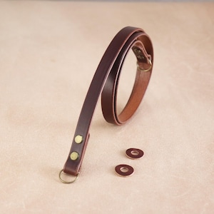 Peut inclure: Une sangle en cuir marron fonc&eacute; avec des &eacute;l&eacute;ments m&eacute;talliques de couleur laiton, dont des rivets et un anneau circulaire. Deux petites pi&egrave;ces de cuir circulaires sont &eacute;galement pr&eacute;sentes. La sangle est probablement pour un sac ou un accessoire.