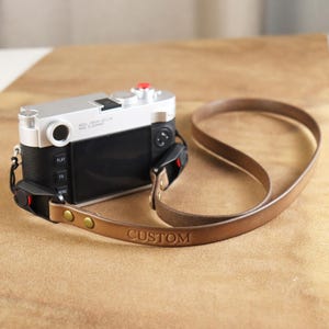 May include: A silver and black camera with a brown leather strap. The strap is embossed with the word "CUSTOM" and is attached to the camera with brass-colored rivets. The camera is on a light brown surface.