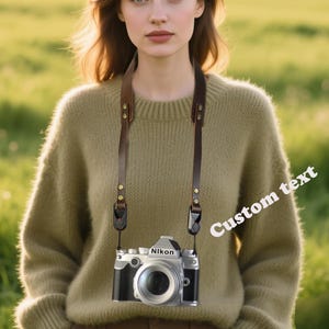 Pode incluir: Uma câmera Nikon vintage com uma alça de couro marrom. A câmera é prateada e preta, com uma lente grande e o logotipo da Nikon. A alça tem rebites cor de latão. O fundo é um campo verde desfocado com o texto "Custom text".