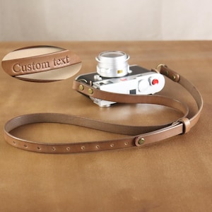 Może przedstawiać: Brązowy skórzany pasek do aparatu z srebrnym aparatem. Pasek ma prosty design z nitami i pętlą. Napis "Custom text" jest wytłoczony na skórzanej etykiecie.