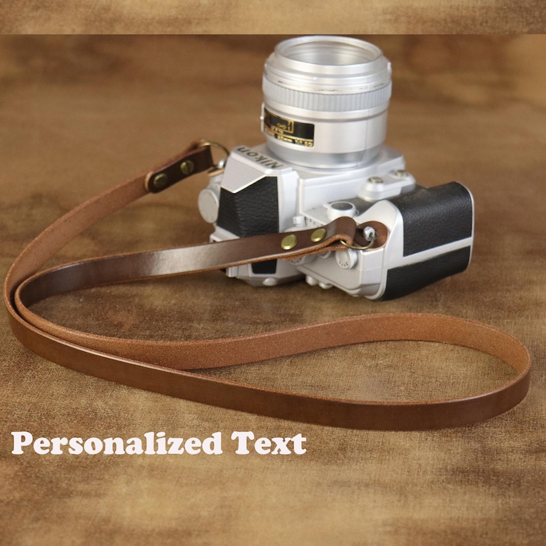 Peut inclure: Une sangle de cam&eacute;ra en cuir marron avec des fixations en laiton est attach&eacute;e &agrave; un appareil photo argent&eacute;. La sangle est longue et fine, avec une boucle &agrave; chaque extr&eacute;mit&eacute;. Le texte "Personalized Text" est au bas de l'image.