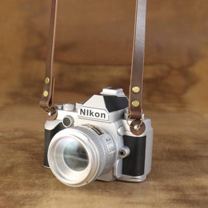 Peut inclure: Un appareil photo Nikon argent&eacute; avec une sangle en cuir marron. L'appareil photo a un grand objectif et des d&eacute;tails noirs. La sangle est fix&eacute;e avec des rivets dor&eacute;s. Le mot "Nikon" est imprim&eacute; sur le corps de l'appareil photo.