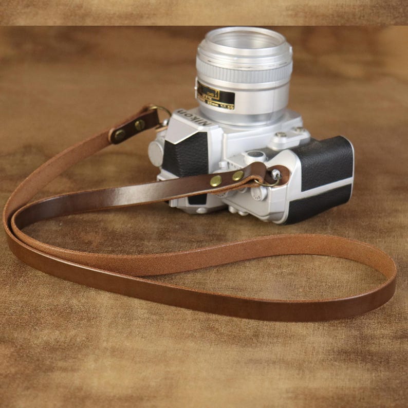 Peut inclure: Une sangle de cam&eacute;ra en cuir marron avec des rivets en laiton est attach&eacute;e &agrave; un appareil photo Nikon argent&eacute;. La sangle est boucl&eacute;e et repose sur une surface marron. L'objectif de l'appareil photo est mis au point.