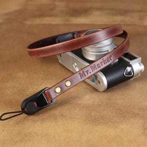 May include: A brown leather camera strap with the text "Mr. Marker" embossed on it. The strap is attached to a black and silver camera. The strap has brass-colored rivets and a black connector.