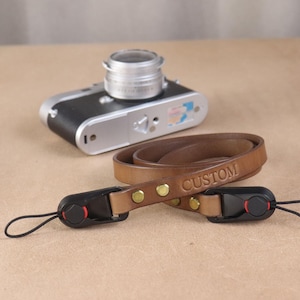 May include: A tan leather camera strap with the word CUSTOM embossed on the surface. The strap features brass rivets and black plastic connectors with thin black loops for attachment. A silver and black camera sits in the background.
