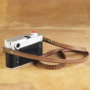 Puede incluir: Cámara plateada con empuñadura de cuero negro y correa de cuero marrón. La correa está sujeta a la cámara con remaches color latón. La cámara tiene un botón rojo en la parte superior.