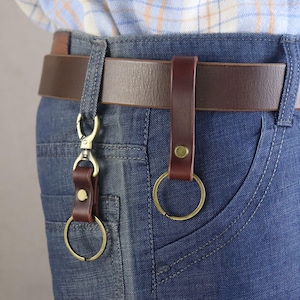 Puede incluir: Primer plano de un llavero de cuero marrón sujeto a un cinturón marrón y unos vaqueros azules. El llavero tiene un anillo color latón y un mosquetón. El cinturón también es de cuero marrón.