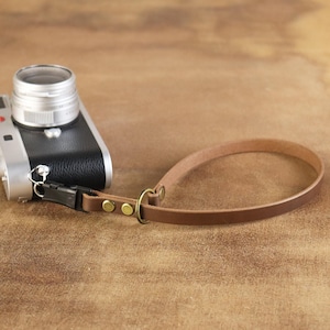 Può includere: Un cinturino da polso per fotocamera in pelle marrone attaccato a una fotocamera argentata e nera. Il cinturino ha un design ad anello con hardware color ottone. L'obiettivo della fotocamera è argentato.
