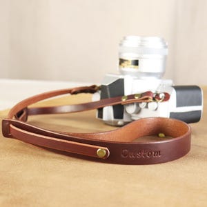 May include: A brown leather camera strap with the word "Custom" embossed on the strap. The strap is attached to a silver camera with a black grip. The strap has brass-coloured rivets and a loop design.