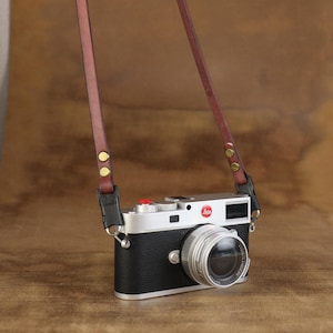 Może przedstawiać: Srebrno-czarny aparat fotograficzny z brązowym skórzanym paskiem. Aparat ma obiektyw i czerwony przycisk na górze. Pasek jest przymocowany do aparatu za pomocą małych metalowych zapięć.