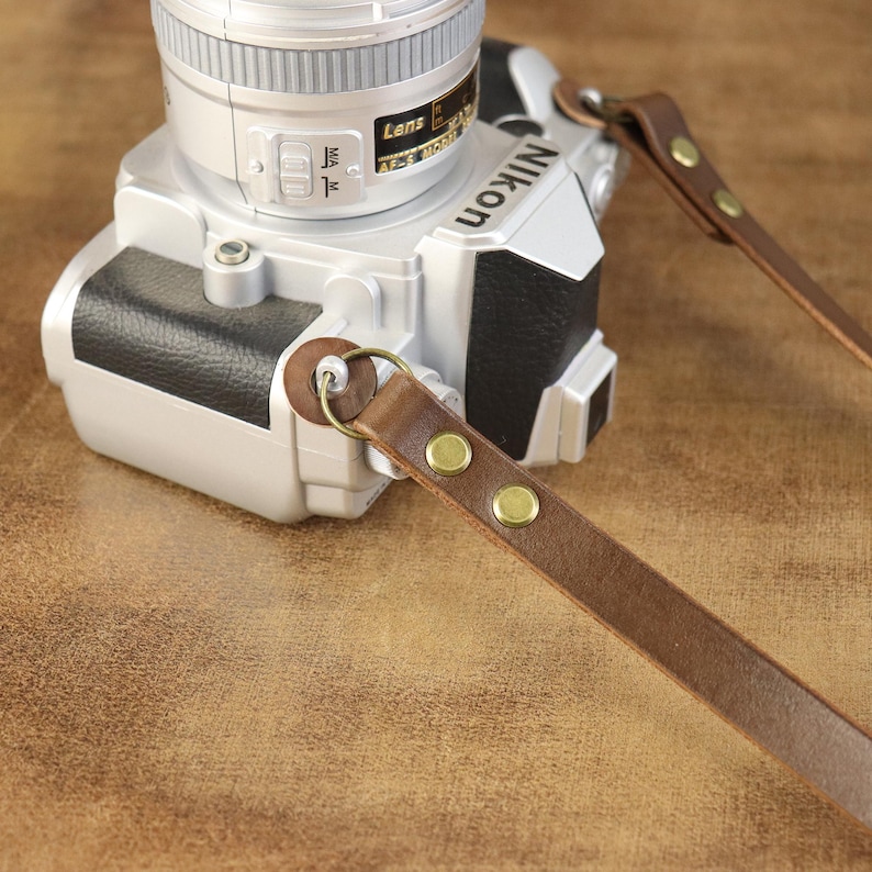 Peut inclure: Une sangle de cam&eacute;ra en cuir marron attach&eacute;e &agrave; un appareil photo Nikon argent&eacute;. La sangle est fix&eacute;e avec des rivets en laiton et un anneau m&eacute;tallique. L'objectif de l'appareil photo est visible.
