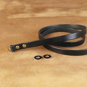 Peut inclure: Sangle d'appareil photo en cuir noir avec rivets en laiton et un anneau dor&eacute;. Deux rondelles en cuir noir et blanc sont &eacute;galement visibles. La sangle est enroul&eacute;e sur une surface marron.