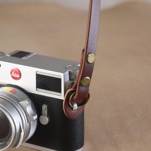 May include: A silver and black camera with a brown leather strap. The camera has a lens and a dial. The strap is attached with brass-colored rivets and a metal ring.