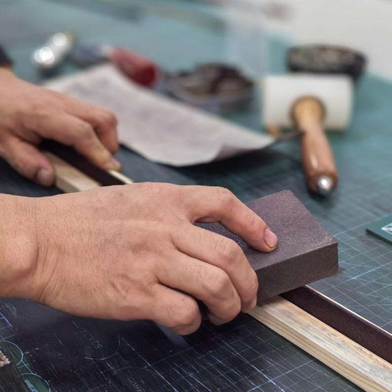Peut inclure: Une personne pon&ccedil;ant un objet en bois avec un bloc de pon&ccedil;age marron. L'objet est en bois clair avec une bande marron fonc&eacute;. Des outils comme un maillet et d'autres articles sont en arri&egrave;re-plan sur une surface verte.