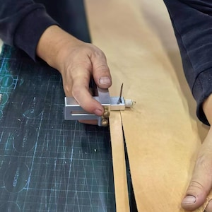 Peut inclure: Une personne utilise un outil de coupe de cuir en m&eacute;tal pour couper une bande de cuir brun clair. L'outil a un corps argent&eacute; avec des accents en laiton et une lame tranchante. Le cuir est sur une surface sombre.