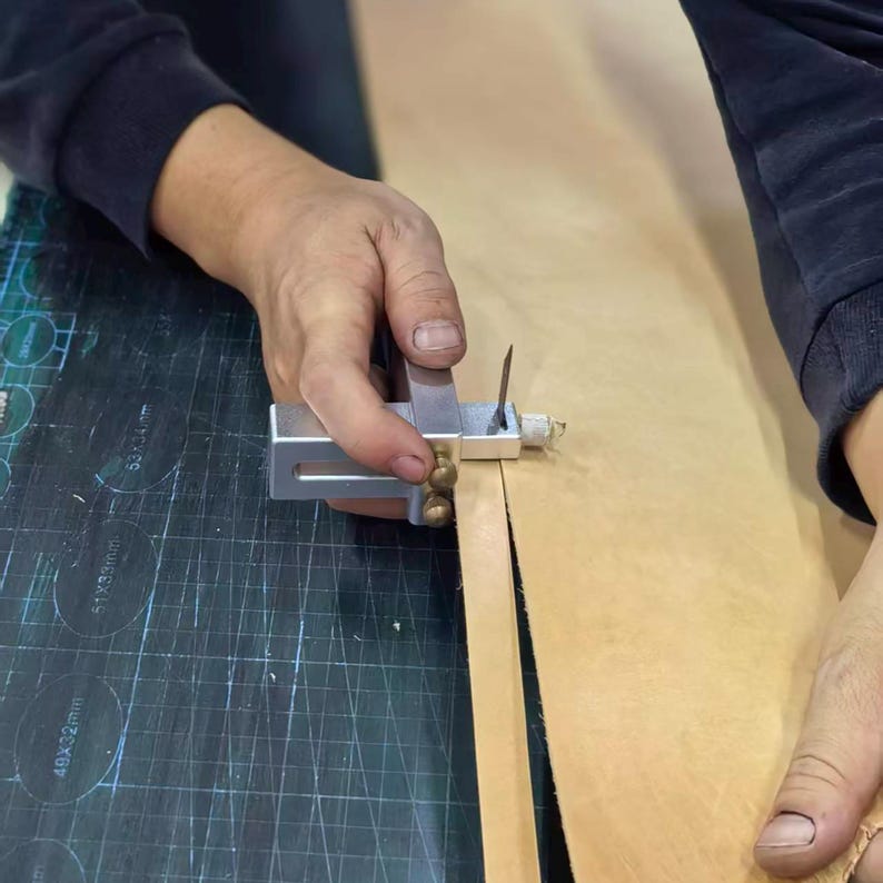 Peut inclure: Une personne utilise un outil de coupe de cuir en m&eacute;tal pour couper une bande de cuir brun clair. L'outil a un corps argent&eacute; avec des accents en laiton et une lame tranchante. Le cuir est sur une surface sombre.