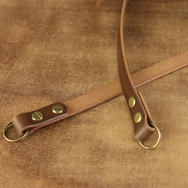 Peut inclure: Gros plan sur des lani&egrave;res en cuir marron avec des ferrures de couleur laiton. Les lani&egrave;res sont fix&eacute;es avec des rivets et comportent des anneaux m&eacute;talliques. Le fond est une surface marron textur&eacute;e, sugg&eacute;rant un produit en cuir.