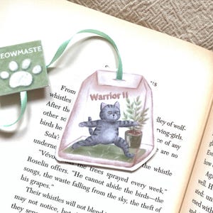 Puede incluir: Un marcapáginas caprichoso con un gato en una postura de yoga, etiquetado como "Warrior II", dentro de un diseño de bolsa de té. El marcapáginas tiene una cinta verde y una pequeña etiqueta cuadrada con el texto "MEOWMASTE" y una huella de pata.