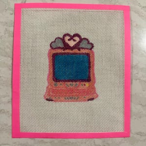 May include: A cross-stitch design featuring a pink retro handheld game console with a blue screen, a heart, and cloud accents. The design is set on a white mesh canvas with a bright pink border.