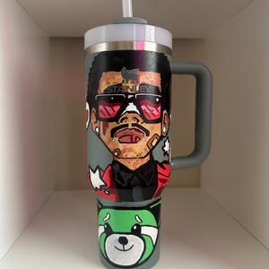 May include: A gray insulated tumbler with a white lid and a gray handle. The tumbler features a hand-painted design of a person wearing sunglasses and a red jacket. The bottom of the tumbler has a green panda design. The word "STANLEY" is visible.