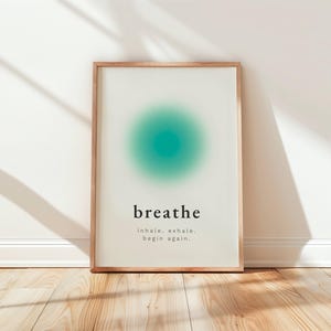Puede incluir: Una impresión artística enmarcada con un marco de madera clara. La impresión presenta la palabra "breathe" en una fuente minimalista, con un diseño circular de color turquesa encima. Debajo, las palabras "inhale, exhale, begin again."