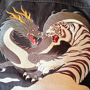 以下が含まれることがあります： 黒いデニムジャケットのクローズアップ。龍と虎が向かい合う刺繍デザインが施されています。龍はグレーと黄色のアクセント、虎は白と茶色の縞模様です。どちらも口を開けています。