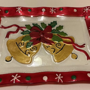 Puede incluir: Una bandeja rectangular de cristal con un diseño festivo de dos campanas doradas atadas con una cinta roja y hojas de acebo verdes. El borde es de color rojo vibrante con copos de nieve blancos y puntos circulares verdes y blancos.