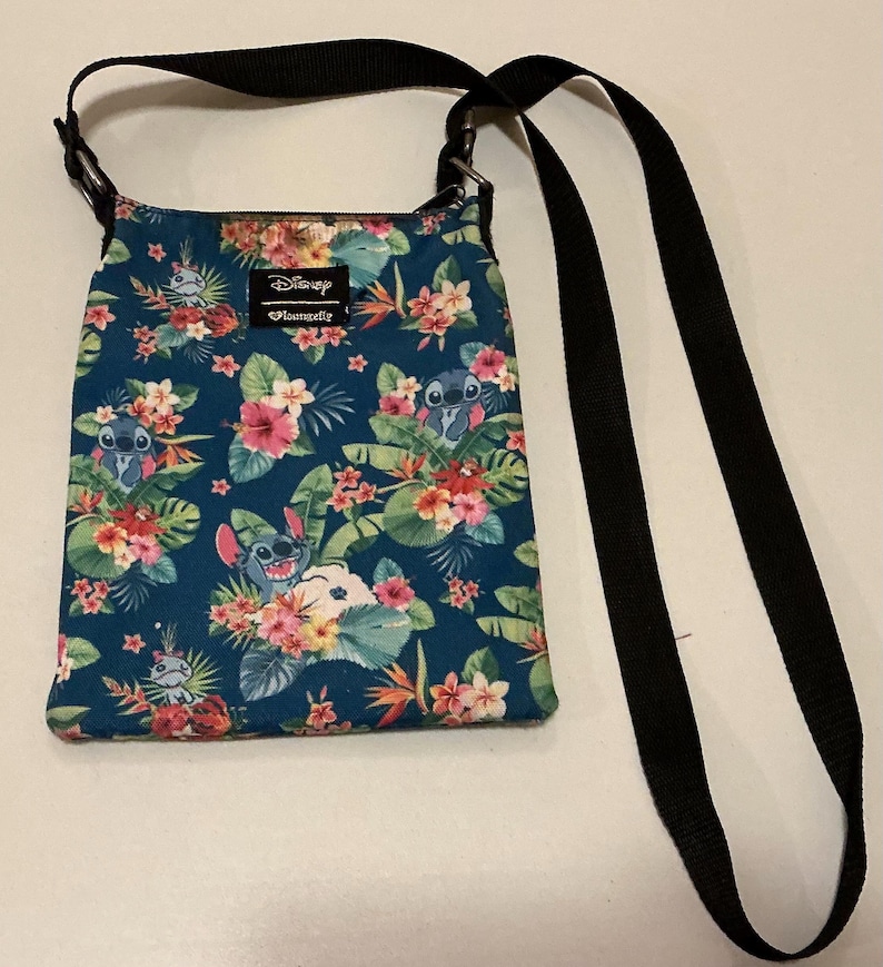 May include: A teal crossbody bag featuring a repeating pattern of the character Stitch from Disney, surrounded by tropical flowers and green leaves. The bag has a black adjustable strap and a rectangular Disney Loungefly brand label.