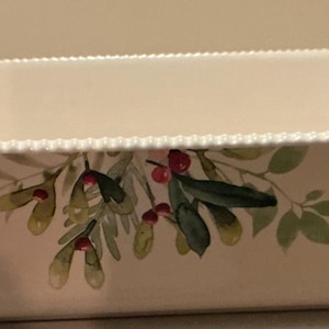 May include: A rectangular cream-coloured ceramic baking dish with integrated handles on both ends. The rim features a delicate beaded texture, and the exterior side is decorated with a painted botanical illustration of green leaves and red berries.