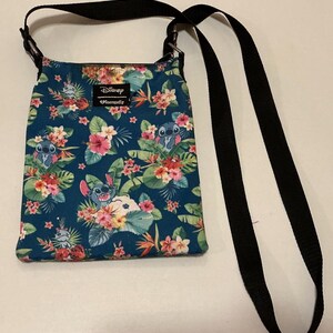 May include: A teal crossbody bag featuring a repeating pattern of the character Stitch from Disney, surrounded by tropical flowers and green leaves. The bag has a black adjustable strap and a rectangular Disney Loungefly brand label.