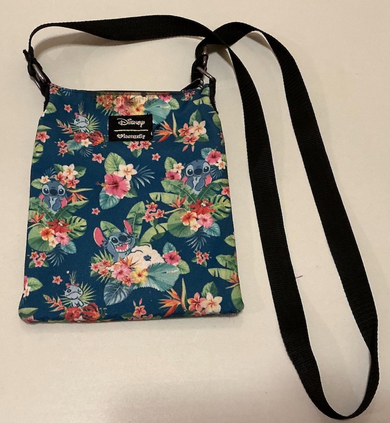May include: A teal crossbody bag featuring a repeating pattern of the character Stitch from Disney, surrounded by tropical flowers and green leaves. The bag has a black adjustable strap and a rectangular Disney Loungefly brand label.