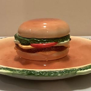 Puede incluir: Una fuente de cerámica con forma de hamburguesa con tapa a juego. La hamburguesa tiene pan, lechuga, tomate, queso y una hamburguesa. El plato es naranja con ribete verde y rojo, ideal para servir aperitivos.