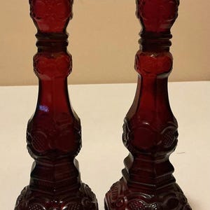 May include: A pair of deep red glass candle holders featuring a pressed pattern with circular and geometric motifs. Each holder has a wide square base, a tapered stem, and a flared top rim, all crafted from translucent ruby-colored glass.