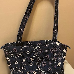 May include: Navy blue quilted tote bag with a floral pattern in white and purple. The bag has two handles and a zipper closure. The bag is approximately 12 inches wide and 10 inches tall.