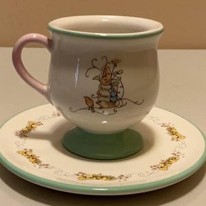 May include: A white ceramic teacup and saucer set with a light green rim and base. The cup features a pink handle and a graphic illustration of Peter Rabbit holding a sign that reads Peter Rabbit. The saucer has a floral border pattern.