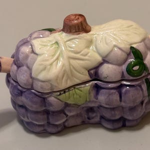 Op de afbeelding: Een keramische sieradendoos in de vorm van een druiventros, met een deksel en twee lepelachtige handvatten. De doos is voornamelijk paars met een cluster van druifachtige vormen. Het deksel heeft een groot bladerontwerp in crème en groen, met een bruine steel.